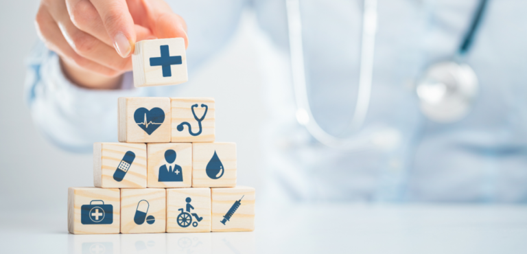 In the background, someone wearing a doctor's coat and stethoscope and in the foreground, they are placing a top block onto a pyramid of wooden blocks, all with different medical icons on them, such as a bandaid, a wheelchair, a drop of blood, etc.