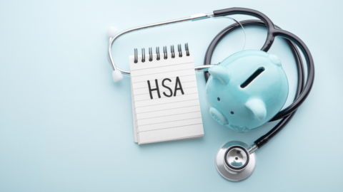 A photo with a light blue background, a notepad that says "HSA" next to a blue piggy bank and a stethoscope wrapped around them