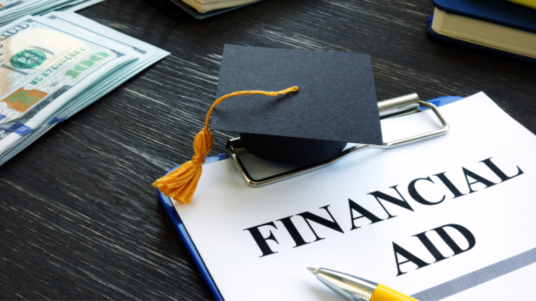 A desktop showing a clipboard with a piece of paper that says "Financial Aid" and on top of it is a graduation cap with some dollar bills in the upper left hand corner.