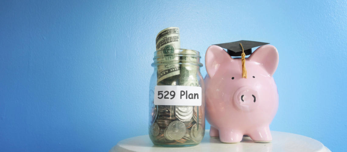 A blue background an in the foreground is a pedestal with a jar that has cash it in and a label that says "529 plan". On the right is a pink porcelain piggy bank that has a graduation cap on it.