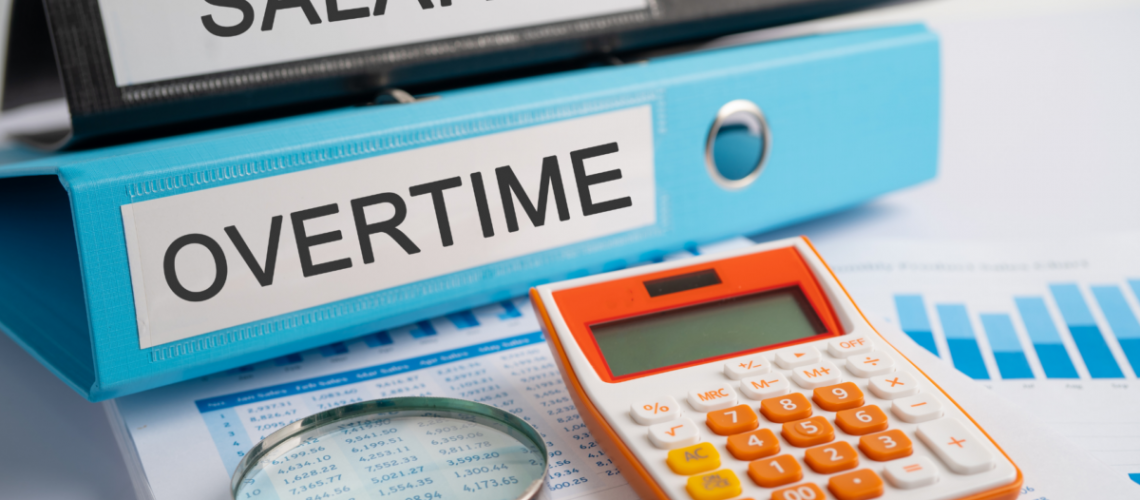 A photo of two binders, one that says "salary" and one that says "overtime". They are stacked on a desk with an orange calculator and a magnifying glass