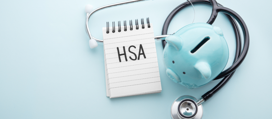 A photo with a light blue background, a notepad that says "HSA" next to a blue piggy bank and a stethoscope wrapped around them