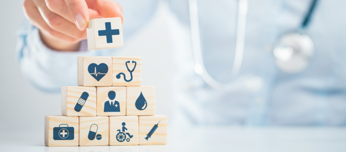 In the background, someone wearing a doctor's coat and stethoscope and in the foreground, they are placing a top block onto a pyramid of wooden blocks, all with different medical icons on them, such as a bandaid, a wheelchair, a drop of blood, etc.