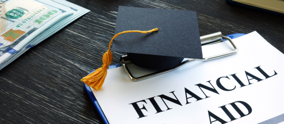 A desktop showing a clipboard with a piece of paper that says "Financial Aid" and on top of it is a graduation cap with some dollar bills in the upper left hand corner.
